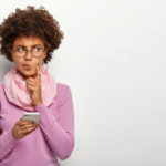 Dark skinned woman with Afro haircut, dressed in violet jumper with scarf, holding mobile phone waiting for invitation to peer review.