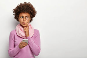 Dark skinned woman with Afro haircut, dressed in violet jumper with scarf, holding mobile phone waiting for invitation to peer review.