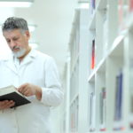 male scientist in white lab coat in library reading book